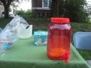 Table covered with a green table cloth, with a lemonade dispenser, cups, pitcher, and cash box on top